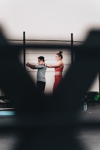 A man and a woman are exercising together, with the woman extending her arms in a workout space. There are blurred foreground elements creating a frame around them. They appear to be using resistance bands, and there is a water bottle on the floor.