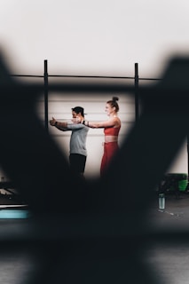 A man and a woman are exercising together, with the woman extending her arms in a workout space. There are blurred foreground elements creating a frame around them. They appear to be using resistance bands, and there is a water bottle on the floor.