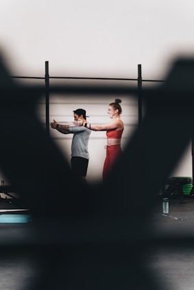 A man and a woman are exercising together, with the woman extending her arms in a workout space. There are blurred foreground elements creating a frame around them. They appear to be using resistance bands, and there is a water bottle on the floor.