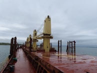 A large cargo ship deck with heavy machinery and cranes stands against an overcast sky. The deck is slightly wet, indicating recent rain. The ocean is visible to the side, with a distant view of land and hills.