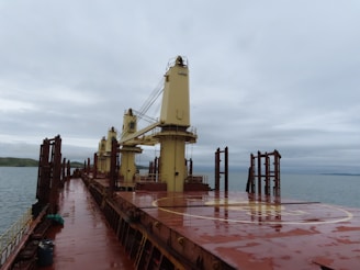 A large cargo ship deck with heavy machinery and cranes stands against an overcast sky. The deck is slightly wet, indicating recent rain. The ocean is visible to the side, with a distant view of land and hills.