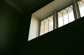 A rectangular window with geometric patterns in the glass panes, allowing natural light to enter a dimly lit room. The contrast between the dark interior and the bright window creates a dramatic effect.