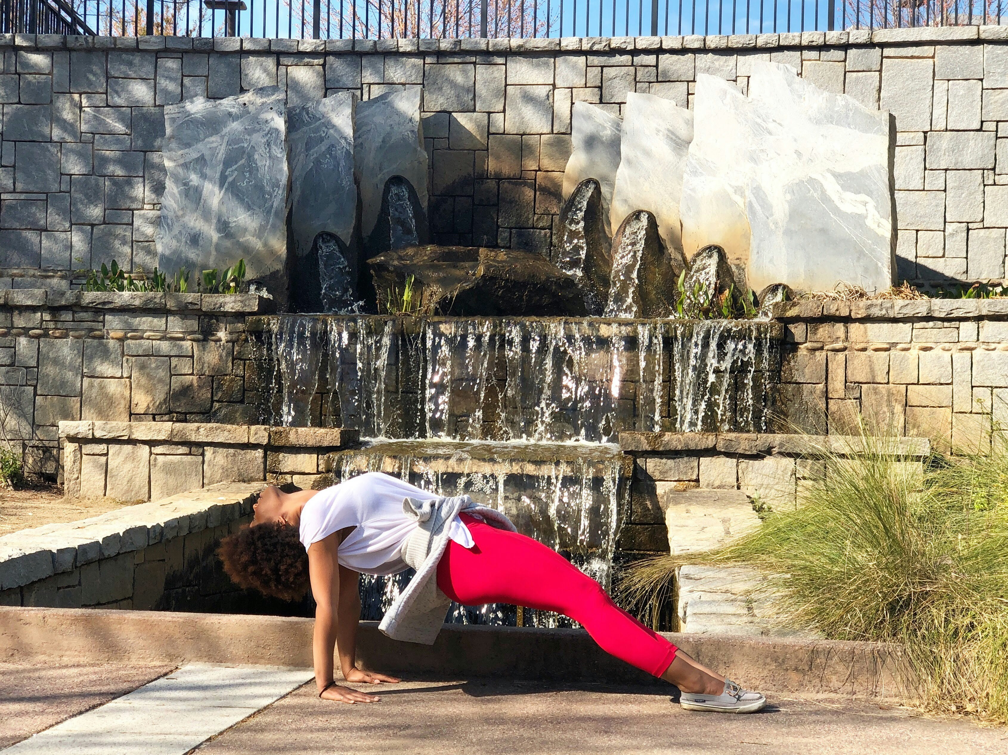 Black woman with an afro practicing Purvottanasana, a yoga pose, in front of a park waterfall. Photo of Christina Mills.