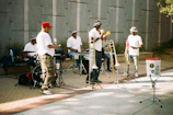 Several musicians are performing outdoors. The group consists of five men wearing mostly white shirts and hats. They are playing a variety of instruments, including drums and trombones, while a man in the center holds maracas. The setting is a paved outdoor area with a gray wall in the background. A white bucket with stickers is placed in front, possibly for collecting donations.