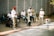 Several musicians are performing outdoors. The group consists of five men wearing mostly white shirts and hats. They are playing a variety of instruments, including drums and trombones, while a man in the center holds maracas. The setting is a paved outdoor area with a gray wall in the background. A white bucket with stickers is placed in front, possibly for collecting donations.