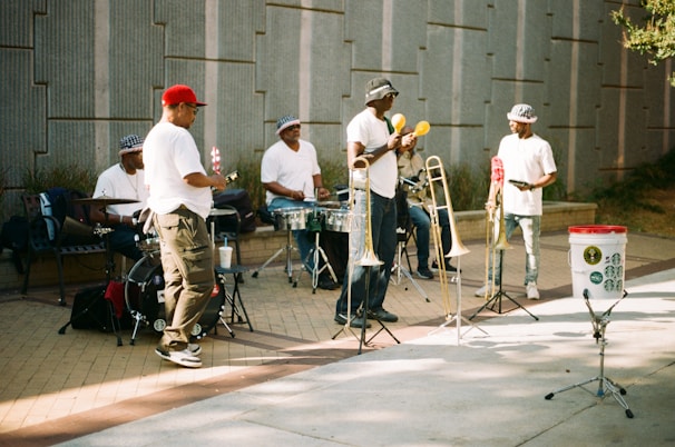 Several musicians are performing outdoors. The group consists of five men wearing mostly white shirts and hats. They are playing a variety of instruments, including drums and trombones, while a man in the center holds maracas. The setting is a paved outdoor area with a gray wall in the background. A white bucket with stickers is placed in front, possibly for collecting donations.