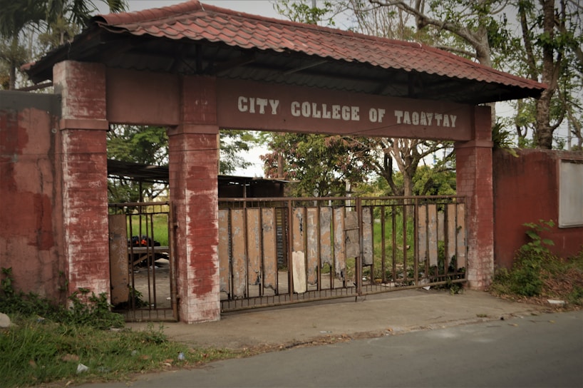 An old entrance gate made of brick and metal with a worn sign displaying 'CITY COLLEGE OF TAGAYTAY'. The gate is partially open, leading to a path surrounded by overgrown greenery. The setting appears to be in a rural or semi-urban area, with trees and grass visible in the background.