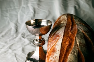 An elegant silver chalice filled with red wine is placed next to a loaf of crusty artisan bread on a textured fabric background. The bread features a deep golden-brown crust with flour dusting.