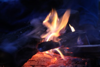 Logs are burning, producing bright flames and glowing embers. The scene is surrounded by darkness, highlighting the vibrant colors of the fire.