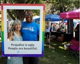 A man and woman are smiling together, framed by a large border with the text, 'Prejudice is ugly. People are beautiful.' They stand in a park setting, with trees and grass visible. Various tents and people in casual clothing are in the background, suggesting an outdoor event or festival.