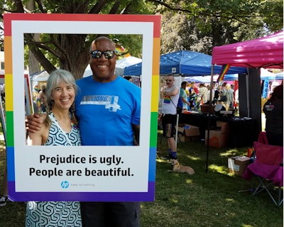 A man and woman are smiling together, framed by a large border with the text, 'Prejudice is ugly. People are beautiful.' They stand in a park setting, with trees and grass visible. Various tents and people in casual clothing are in the background, suggesting an outdoor event or festival.