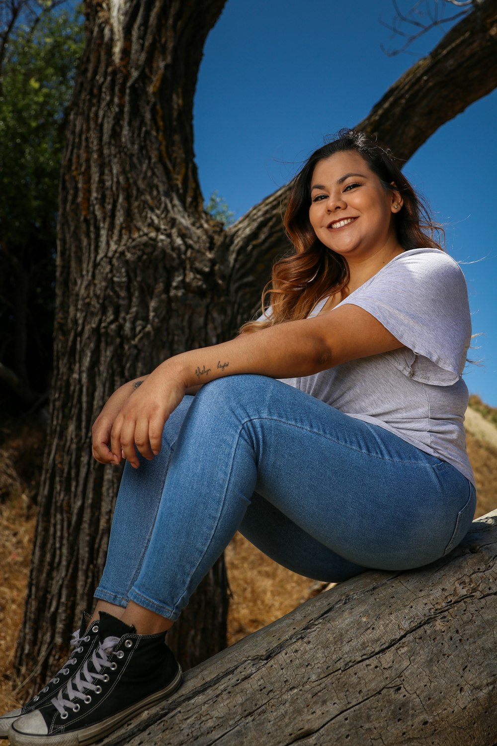 Mujer con camisa gris y jeans de mezclilla azul sentada en el tronco de un árbol marrón durante el día