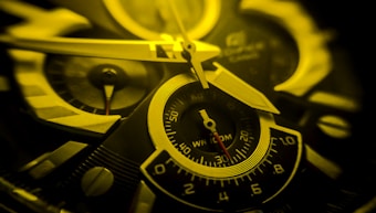 A close-up view of a watch face highlighting the intricate details of the clock hands and dials. The image is predominantly colored in shades of yellow and black, emphasizing the precision and design of the watch mechanism.