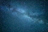 A dense cluster of stars and cosmic dust forming a segment of the Milky Way galaxy stretches diagonally across a dark night sky. Countless stars with varying brightness are scattered throughout the image.