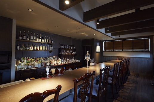 A dimly lit bar with a wooden countertop and a row of intricate wooden chairs. The back wall features shelves lined with various bottles of liquor. Glasses and drinks are arranged neatly on the bar, including a couple of cocktails. The space has a warm, cozy atmosphere with dark wooden paneling and overhead ceiling beams.