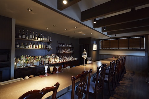 A dimly lit bar with a wooden countertop and a row of intricate wooden chairs. The back wall features shelves lined with various bottles of liquor. Glasses and drinks are arranged neatly on the bar, including a couple of cocktails. The space has a warm, cozy atmosphere with dark wooden paneling and overhead ceiling beams.