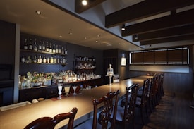 A dimly lit bar with a wooden countertop and a row of intricate wooden chairs. The back wall features shelves lined with various bottles of liquor. Glasses and drinks are arranged neatly on the bar, including a couple of cocktails. The space has a warm, cozy atmosphere with dark wooden paneling and overhead ceiling beams.