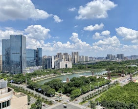 The image depicts an urban area with high-rise buildings and a green landscape. A river flows through the center, surrounded by lush trees and parks. The sky is clear, with scattered fluffy clouds.