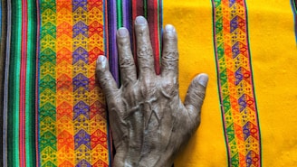 A wrinkled hand is placed on a colorful textile with intricate patterns. The textile features vibrant colors like yellow, orange, purple, and green in various geometric designs.