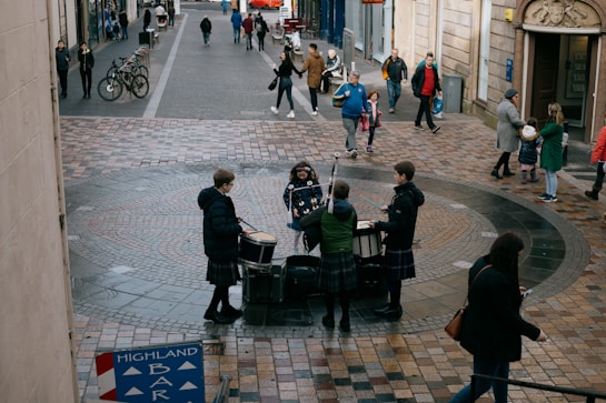 A group of musicians dressed in traditional attire perform with drums and a bagpipe in a cobblestone courtyard. People are walking by and a few are stopping to watch. Bicycles are parked at the side and there are several pedestrians along the street.