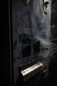 A close-up view of a worn, vintage vending machine with partial text reading 'einzigartig' on its surface. Smoking wisps trail across the machine, adding a mysterious and somewhat eerie effect. The machine appears old and slightly rusted, with dark blue and metallic tones.