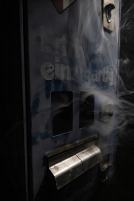 A close-up view of a worn, vintage vending machine with partial text reading 'einzigartig' on its surface. Smoking wisps trail across the machine, adding a mysterious and somewhat eerie effect. The machine appears old and slightly rusted, with dark blue and metallic tones.