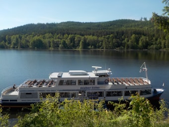 A large white passenger boat is docked by a calm, expansive lake surrounded by lush green forest. The boat has multiple rows of seating visible on the upper deck, and its name, 'Adalbert Stifter,' is painted on the side. The sky is clear, and the sunlight highlights the vibrant greenery and the serene waters.