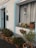 A charming exterior of a house is adorned with various potted plants. Several terracotta pots with lush greenery line the base of a white wall with blue trim. A window sill holds more plants, including blooming flowers. A hanging plant in a blue pot adds a vertical element. An old-fashioned mailbox and a house number are visible near a teal door.