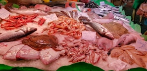A seafood display at a market featuring a variety of fish and shellfish. The arrangement includes red shrimp, whole fish like sea bass, cuttlefish, and possibly flatfish. Ice is used as a base to keep the seafood fresh, and there are handwritten price tags indicating different prices for each item.