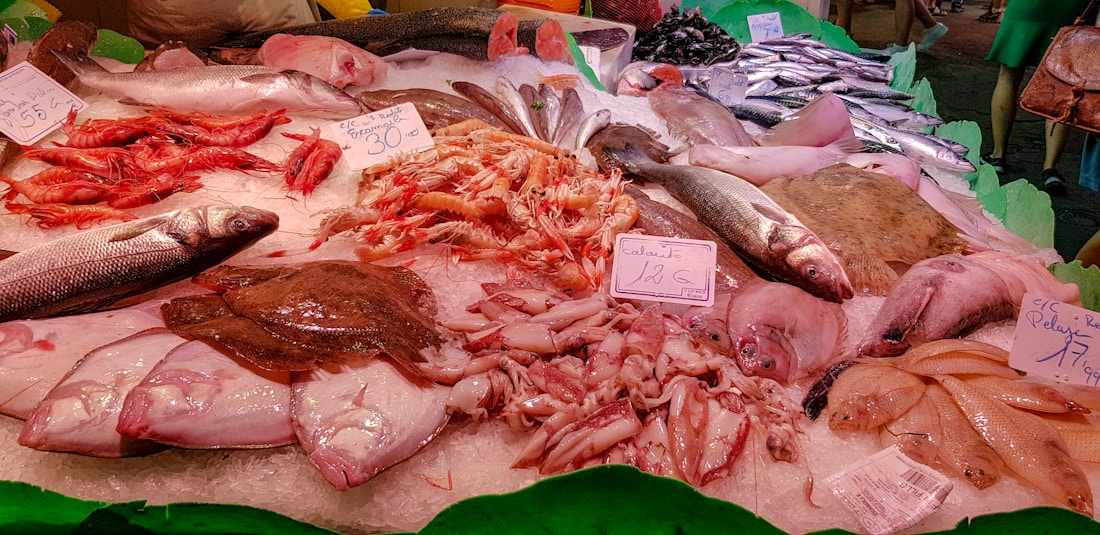 A seafood display at a market featuring a variety of fish and shellfish. The arrangement includes red shrimp, whole fish like sea bass, cuttlefish, and possibly flatfish. Ice is used as a base to keep the seafood fresh, and there are handwritten price tags indicating different prices for each item.