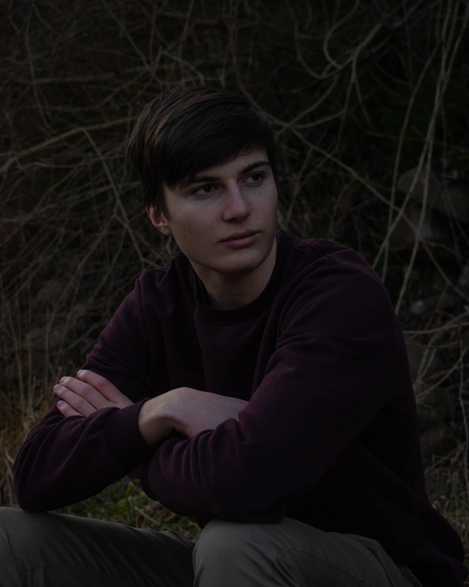 Young man sits on ground, wearing purple sweater, gazing off into darkness surrounded by trees