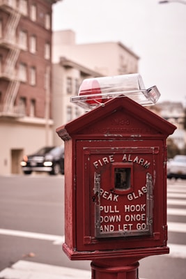 An old-fashioned red fire alarm box stands on a street corner against the backdrop of urban buildings. The alarm box features instructions that read 'Break Glass, Pull Hook Down Once and Let Go,' and is topped with a glass cover. The background is slightly blurred, showing a brick building and a parked car.