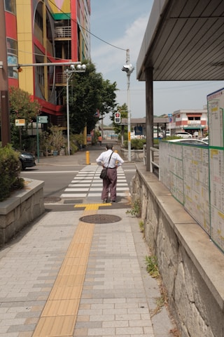 A person stands at a crosswalk on a city street, facing away. The area features a colorful building with geometric designs, overhead traffic lights, and a sidewalk with tactile paving tiles. A map or information board is visible on the right side.
