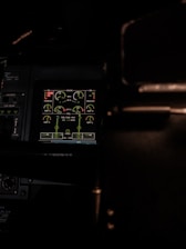 A dimly lit cockpit control panel with various gauges and digital displays showing information related to engine performance, oil levels, and fuel indicators. The screen is highlighted in green, surrounded by dark panels and some buttons.