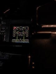 A dimly lit cockpit control panel with various gauges and digital displays showing information related to engine performance, oil levels, and fuel indicators. The screen is highlighted in green, surrounded by dark panels and some buttons.