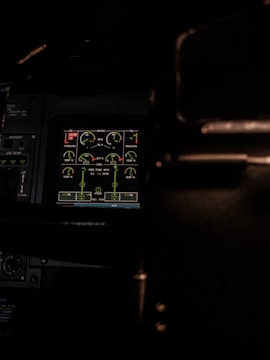 A dimly lit cockpit control panel with various gauges and digital displays showing information related to engine performance, oil levels, and fuel indicators. The screen is highlighted in green, surrounded by dark panels and some buttons.