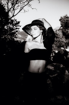 A stylish woman stands confidently outdoors. She wears a wide-brimmed hat and a sleeveless top that reveals her midriff. The lighting creates strong contrasts, highlighting her face and upper body against a darker background of trees and foliage.