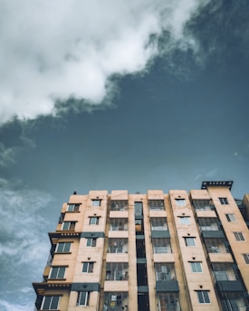 A multi-story residential building with a beige facade and several windows is viewed from a low angle. The sky above is mostly overcast with some patches of clearer, darker sky visible near the edges. The building has a rugged, urban appearance with visible balconies and some wires.