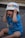 A young child wearing a blue shirt with the words 'VIP BABY' is playfully holding a decorated plastic bowl on their head while sitting on a tiled floor. The child's expression is joyful and engaged.