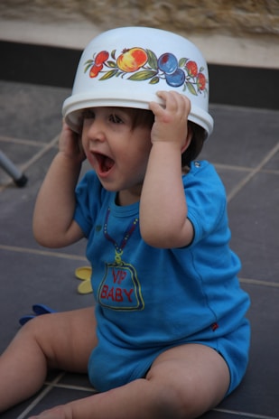 A young child wearing a blue shirt with the words 'VIP BABY' is playfully holding a decorated plastic bowl on their head while sitting on a tiled floor. The child's expression is joyful and engaged.