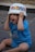 A young child wearing a blue shirt with the words 'VIP BABY' is playfully holding a decorated plastic bowl on their head while sitting on a tiled floor. The child's expression is joyful and engaged.
