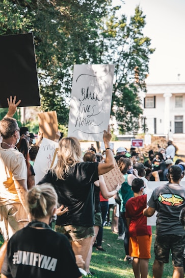 A diverse group of people gathered outdoors holding signs and participating in a protest. The setting is a grassy area with trees and buildings in the background. The signs feature messages promoting social justice.
