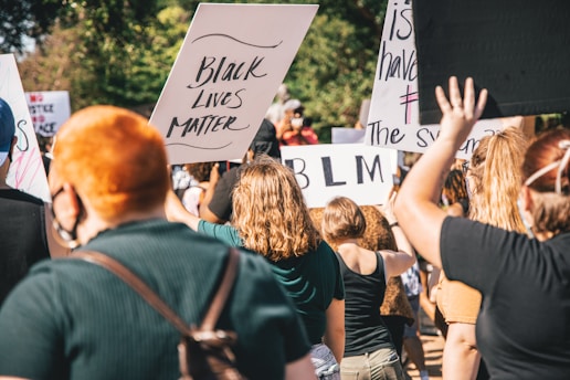 A gathering of people holding protest signs with phrases like 'Black Lives Matter' and 'BLM'. The crowd appears to be diverse in age and gender, with individuals wearing casual summer clothing. Many participants have their backs to the camera and are raising their signs and hands. The setting suggests a public outdoor space, with trees visible in the background.