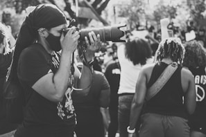 A person wearing a mask and dark clothing is taking a photograph with a large camera lens. They are surrounded by a crowd of people, some of whom have raised fists. The scene appears candid and dynamic with elements of a public gathering or protest.