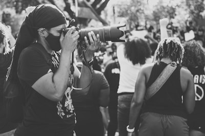 A person wearing a mask and dark clothing is taking a photograph with a large camera lens. They are surrounded by a crowd of people, some of whom have raised fists. The scene appears candid and dynamic with elements of a public gathering or protest.
