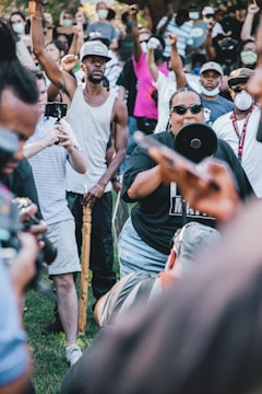 A group of people gathered outdoors, many raising their fists in the air, conveying a sense of unity and protest. One person prominently holds a megaphone, suggesting leadership or the act of addressing the crowd. Individuals are dressed casually, with some wearing masks and sunglasses, indicative of a contemporary setting possibly during a health crisis or pandemic.