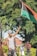 A man is holding up a Pan-African flag in an outdoor setting with trees in the background. He is wearing a grey tank top and a cap, while other individuals stand around, some wearing masks.