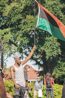 A man is holding up a Pan-African flag in an outdoor setting with trees in the background. He is wearing a grey tank top and a cap, while other individuals stand around, some wearing masks.
