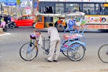 A vibrant rickshaw decorated with colorful ornaments stands on a busy city street. A man is leaning on the rickshaw, which is painted in vivid blues and has intricate patterns. In the background, a crowded bus and a red car can be seen, along with people on motorcycles, indicating a bustling urban environment.