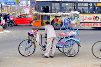 A vibrant rickshaw decorated with colorful ornaments stands on a busy city street. A man is leaning on the rickshaw, which is painted in vivid blues and has intricate patterns. In the background, a crowded bus and a red car can be seen, along with people on motorcycles, indicating a bustling urban environment.